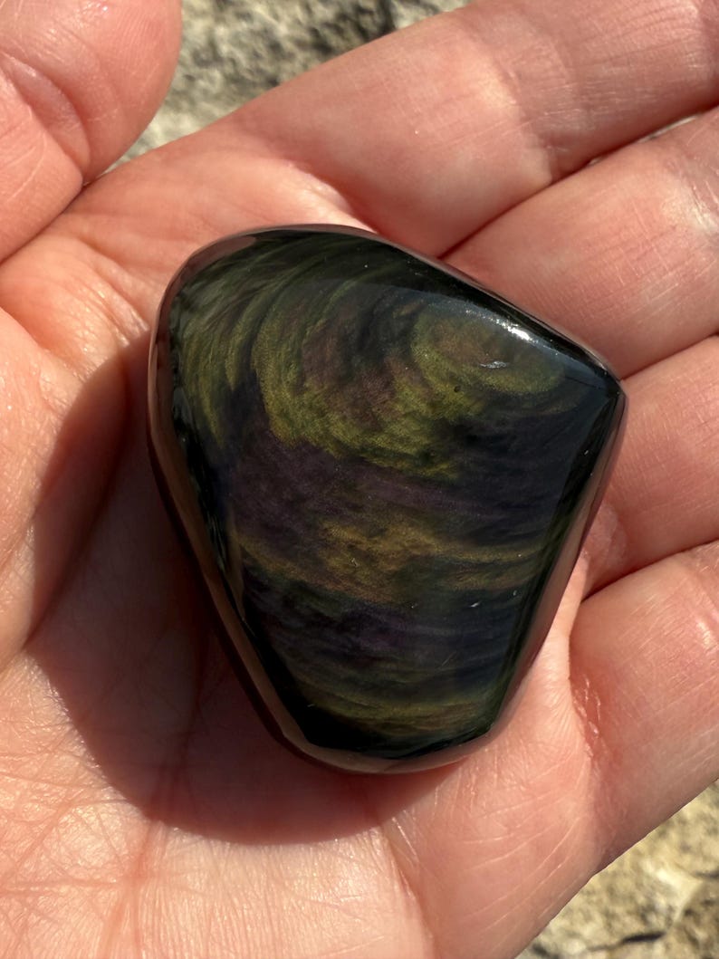 Puede incluir: Una piedra de obsidiana pulida con patrones arremolinados de verde, dorado y p&uacute;rpura. La piedra es lisa y redondeada, sostenida en una mano abierta, y refleja la luz, mostrando sus colores y texturas naturales.