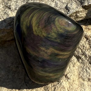 Puede incluir: Una piedra de obsidiana pulida con patrones arremolinados de verde, dorado y p&uacute;rpura. La forma lisa y redondeada de la piedra contrasta con la superficie rugosa y texturizada sobre la que descansa. Los colores de la piedra cambian con la luz.