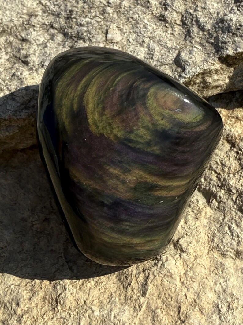 Puede incluir: Una piedra de obsidiana pulida con patrones arremolinados de verde, dorado y p&uacute;rpura. La forma lisa y redondeada de la piedra contrasta con la superficie rugosa y texturizada sobre la que descansa. Los colores de la piedra cambian con la luz.