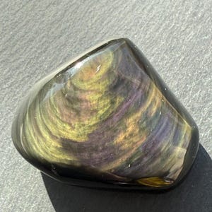Puede incluir: Una piedra preciosa de obsidiana pulida con patrones arremolinados de oro, p&uacute;rpura y verde. La piedra tiene una forma suave y redondeada y un acabado brillante, que refleja la luz. La piedra natural es un art&iacute;culo decorativo &uacute;nico.