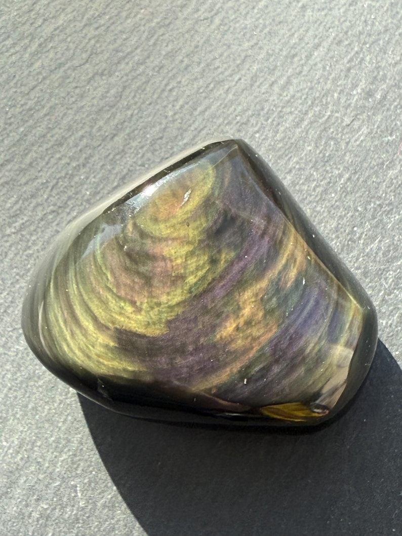 Puede incluir: Una piedra preciosa de obsidiana pulida con patrones arremolinados de oro, p&uacute;rpura y verde. La piedra tiene una forma suave y redondeada y un acabado brillante, que refleja la luz. La piedra natural es un art&iacute;culo decorativo &uacute;nico.