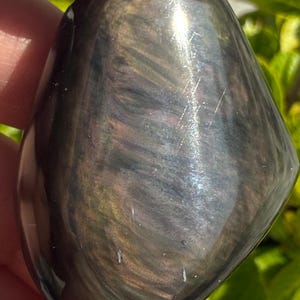 Puede incluir: Una piedra preciosa pulida en forma de l&aacute;grima con una superficie oscura e iridiscente. La piedra muestra patrones arremolinados de oro, plata y negro, capturando la luz para crear un efecto brillante. La piedra preciosa se sostiene en una mano, con follaje verde en el fondo.