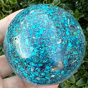 May include: A spherical gemstone, predominantly turquoise and teal, with a mosaic pattern set in a dark base. The polished sphere is held in a hand, with green foliage in the background.