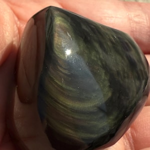 Puede incluir: Una piedra de obsidiana pulida y redondeada, con una superficie oscura y reflectante. Presenta remolinos iridiscentes de oro y verde, creando un efecto brillante. La superficie lisa de la piedra se destaca por la luz natural.