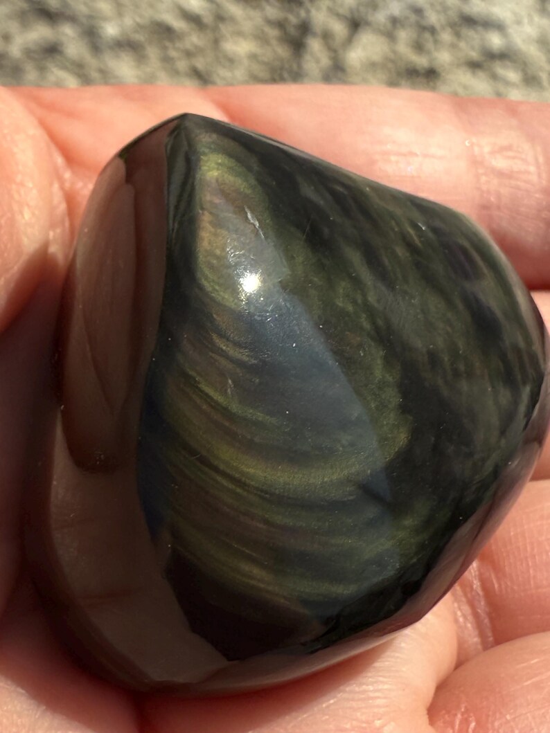 Puede incluir: Una piedra de obsidiana pulida y redondeada, con una superficie oscura y reflectante. Presenta remolinos iridiscentes de oro y verde, creando un efecto brillante. La superficie lisa de la piedra se destaca por la luz natural.