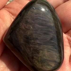 Puede incluir: Una piedra de obsidiana pulida e iridiscente con una forma suave y redondeada. La piedra muestra una mezcla de colores verde oscuro, p&uacute;rpura y negro, con un acabado brillante. La piedra preciosa se sostiene en una mano.