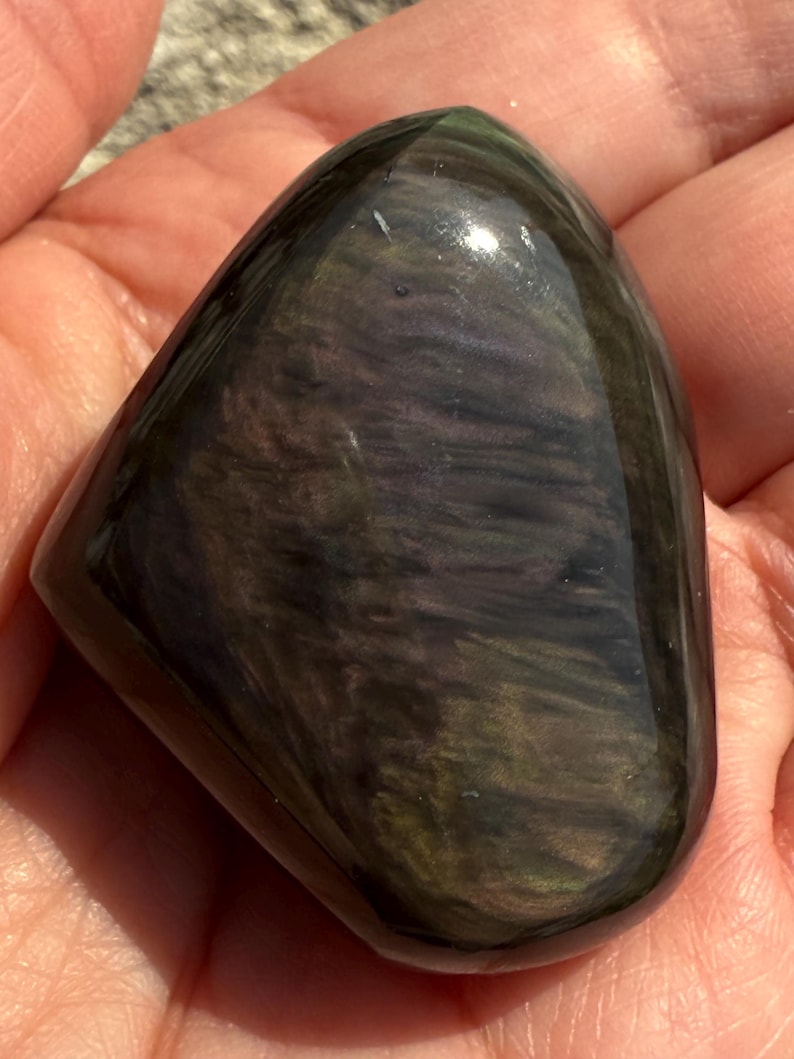 Puede incluir: Una piedra de obsidiana pulida e iridiscente con una forma suave y redondeada. La piedra muestra una mezcla de colores verde oscuro, p&uacute;rpura y negro, con un acabado brillante. La piedra preciosa se sostiene en una mano.