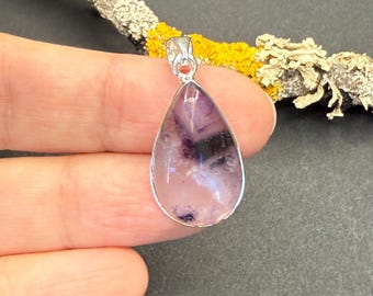 May include: A teardrop-shaped pendant with a silver-colored bail. The pendant features a polished, translucent stone with shades of purple, white, and black. The stone is set in a silver-colored bezel. The background includes a branch with yellow and white lichen.