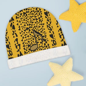 May include: A yellow beanie with a black leopard print and a musical note graphic. The hat has a white cuff and is next to two yellow star-shaped decorative items. The beanie is a stylish accessory.