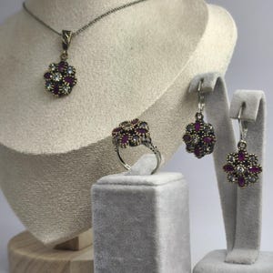 May include: A jewelry set featuring a necklace, ring, and earrings. Each piece has a floral design with dark red and clear gemstones set in a silver-toned metal. The jewelry is displayed on a mannequin bust and velvet stands.