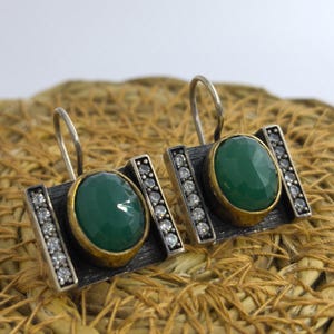 May include: A pair of silver earrings with a rectangular design. Each earring features a large, round, green gemstone set in a gold-colored bezel. The rectangular frame is accented with small, clear gemstones.
