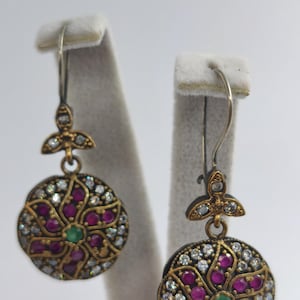 May include: A pair of ornate earrings with a floral design. Each earring features a circular pendant with a gold-colored frame, encrusted with small, round, red and white gemstones. A green gemstone is at the center.