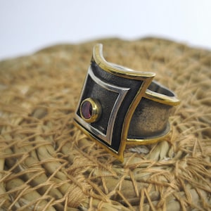 May include: A silver ring with a geometric design, featuring a black center and gold trim. A small, round, red gemstone is set in the center of the design. The ring is displayed on a woven, tan surface.