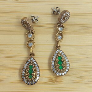 Puede incluir: Un par de pendientes colgantes de tono dorado con forma de lágrima. Los pendientes están adornados con piedras preciosas transparentes y verdes.