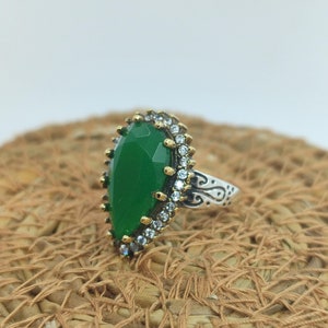 May include: A silver ring featuring a large green gemstone surrounded by smaller clear stones. The ring band has an intricate design.