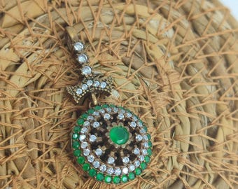 May include: A silver pendant with a round green stone surrounded by clear and green crystals. The pendant has a detailed design and is hanging from a silver chain.