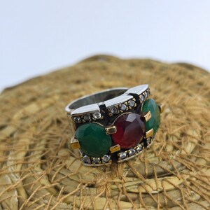 Puede incluir: Un anillo plateado con tres piedras preciosas grandes: una piedra roja central flanqueada por dos piedras verdes. El anillo está adornado con pequeñas piedras transparentes y detalles dorados. El anillo está sobre una superficie tejida y texturizada.