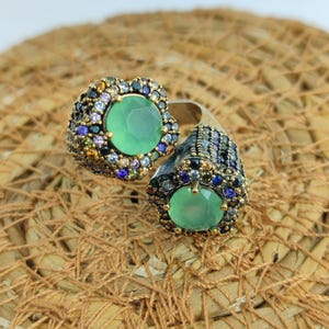 May include: A silver ring with two green gemstones surrounded by colorful rhinestones. The ring is designed in a floral shape.