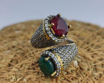 May include: Two ornate rings with large gemstones. One ring features a deep red gemstone, while the other showcases a vibrant green stone. Both rings have a silver band encrusted with small, sparkling stones and gold accents around the gemstones.