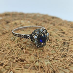 Puede incluir: Un anillo de plata con una piedra preciosa azul rodeada de piedras blancas más pequeñas. El anillo tiene una banda detallada con un diseño floral.