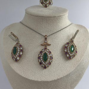 May include: A jewellery set comprising a necklace, earrings, and a ring. Each item features an oval green stone encircled by small white and red stones. The set is displayed on a mannequin head against a white backdrop.