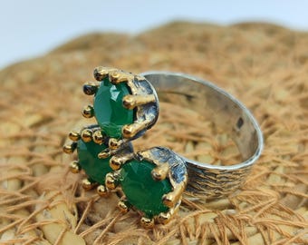 May include: A silver ring with three green gemstones set in gold. The ring has a textured band and is designed to be worn on the knuckle.