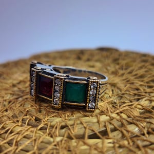 Puede incluir: Un anillo plateado con dos engastes rectangulares, uno rojo y otro verde, cada uno enmarcado con oro y pequeñas piedras transparentes. El anillo está sobre un fondo texturizado y tejido.