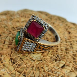 Puede incluir: Dos anillos ornamentados con piedras cuadradas. Uno presenta una gema roja vibrante, el otro una gema verde. Ambos anillos tienen una banda plateada con detalles dorados y están incrustados con pequeñas piedras transparentes alrededor de las gemas.