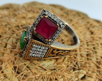 May include: Two ornate rings with square-cut gemstones. One ring features a vibrant red gemstone, the other a green gemstone. Both rings have a silver band with gold accents and are encrusted with small, clear stones around the gemstones.