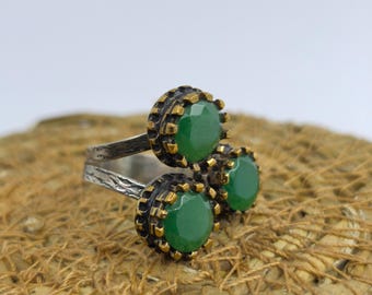 May include: A silver ring with three green gemstone settings. Each setting has a dark, textured border with gold accents. The ring is displayed on a woven, brown surface.