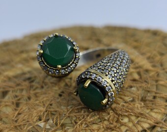 May include: Two ornate rings with large, round, emerald-green gemstones. One ring features a halo of small, clear stones around the green gem. The ring bands are silver-toned, and the settings have gold accents. The rings are displayed on a textured, brown surface.