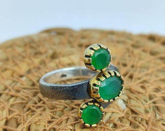 May include: A silver ring with three green gemstones. The gemstones are set in gold-colored bezels. The ring has a textured band and a unique, eye-catching design. The gemstones are oval and round.