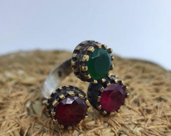 May include: A silver ring with three gemstones. The ring features a green gemstone and two red gemstones, each set in a decorative gold-toned bezel. The ring is displayed on a woven, textured surface.