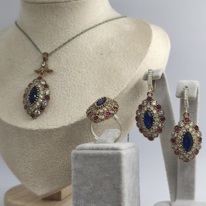 May include: A jewelry set featuring a necklace, ring, and earrings. The pieces have a central blue stone surrounded by smaller white and red stones. The necklace and earrings are suspended from gold-colored settings, while the ring sits on a silver band.