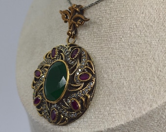 May include: A gold-toned pendant necklace with an oval green gemstone center. The pendant is adorned with red gemstones and small clear stones. The necklace is displayed on a neutral-colored mannequin.