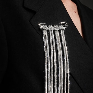 May include: A silver brooch with dangling crystal strands is pinned to a black blazer. The brooch features a row of crystals above the cascading strands, creating a sparkling, elegant accessory. The blazer's fabric is smooth and dark.