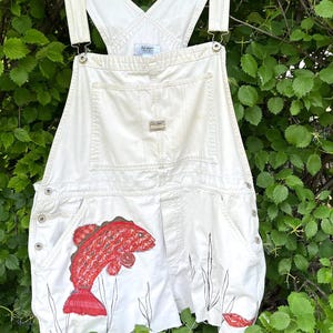 May include: White denim overalls with a red fish embroidery on the front pocket. The overalls have adjustable straps, metal hardware, and a classic bib pocket design. The Old Navy brand is visible on the bib.