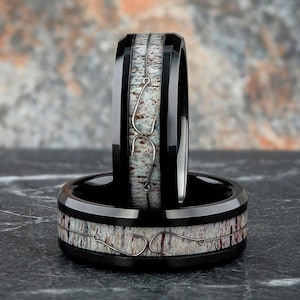 May include: Two black rings with a white and brown speckled inlay. Each ring features a silver fishing hook design. The rings are stacked, with one ring in the foreground and the other standing upright behind it.