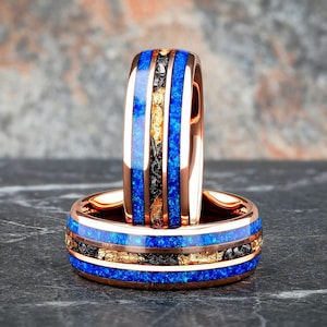 May include: Two rose gold rings with a blue, black, and gold inlay design. The rings have a polished finish and are set against a dark gray surface. The rings are a unique piece of jewelry.