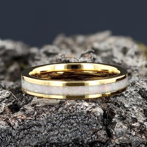 May include: A gold ring with a thin band of white opal inlay. The ring is sitting on a piece of rough, gray bark.