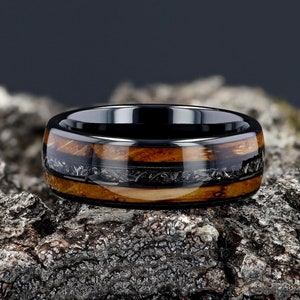 May include: A black ceramic ring with a band of wood and meteorite inlay. The ring has a polished finish and is set on a rough, textured surface.