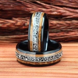 May include: Two black rings with a wood grain center band and a silver filigree design. The rings are set on a wooden surface with a blurred background of dried plants.