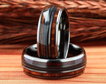 Puede incluir: Dos anillos de carburo de tungsteno negro con incrustaciones de madera. Los anillos tienen un diseño acanalado y están apilados uno encima del otro.