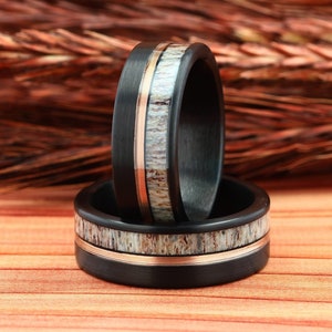 Puede incluir: Dos anillos de fibra de carbono negra con una banda fina de oro rosa y una incrustaci&oacute;n de asta blanca. Los anillos est&aacute;n apilados uno encima del otro.
