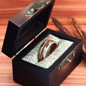 May include: A wooden ring box with a brown and black finish, open to reveal a gold and copper ring with a black band.