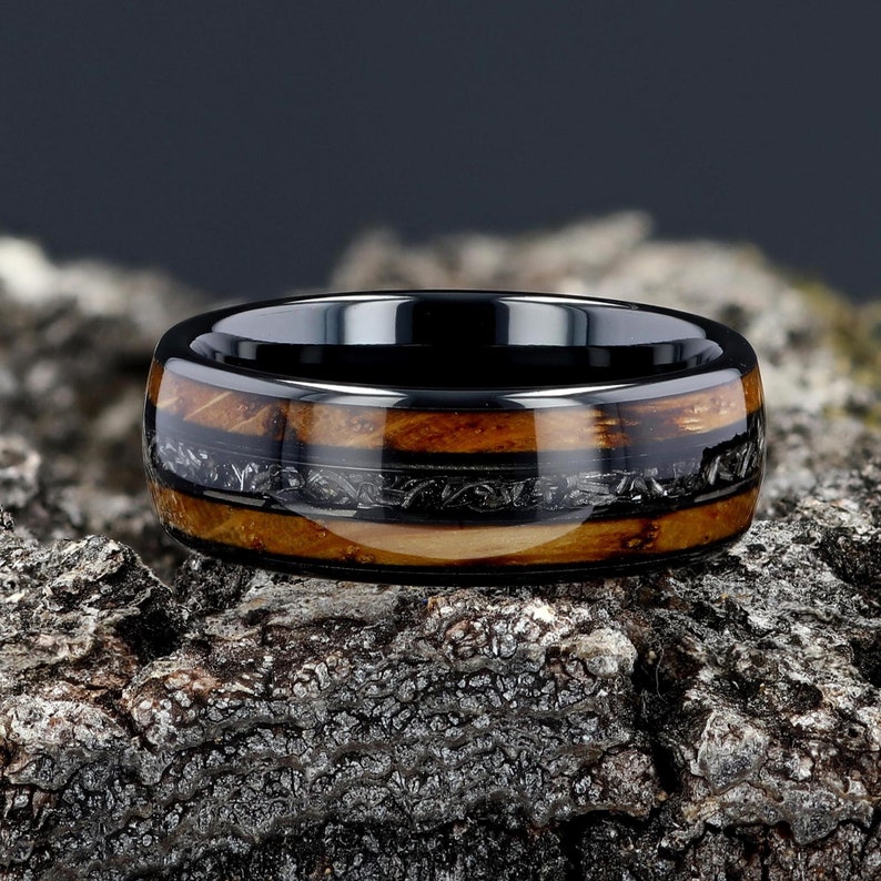 May include: A black ceramic ring with a band of wood and meteorite inlay. The ring has a polished finish and is set on a rough, textured surface.