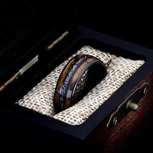 May include: A black tungsten carbide ring with a brown wood inlay and a silver stripe. The ring is in a wooden box with a beige fabric lining.