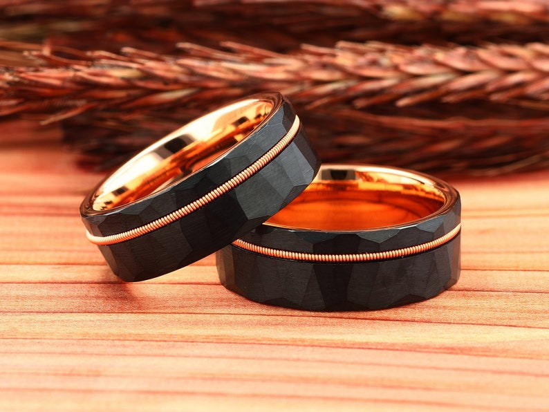 Puede incluir: Dos anillos de carburo de tungsteno negro con una incrustaci&oacute;n de oro rosa. Los anillos tienen un acabado martillado y una fina banda de oro rosa alrededor del centro.