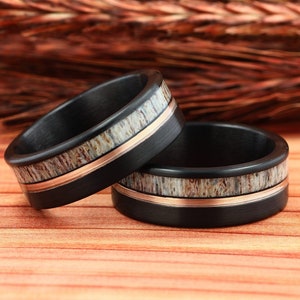 Puede incluir: Dos anillos de boda de fibra de carbono negra con una banda fina de oro rosa y una tira de incrustaci&oacute;n de asta.