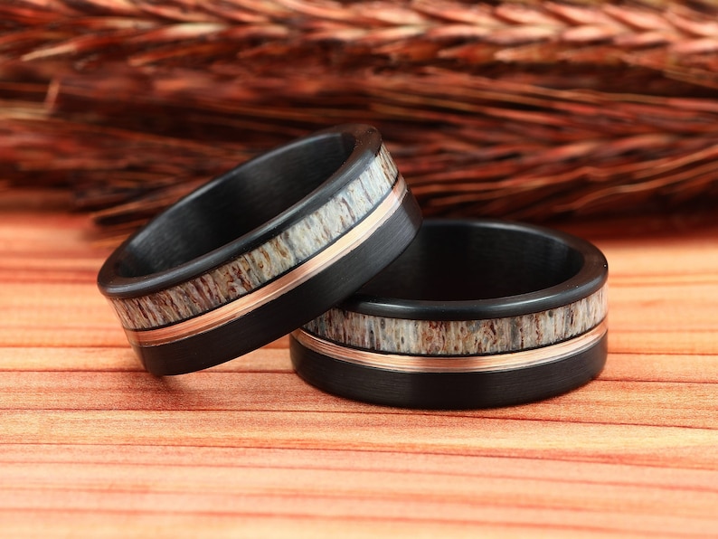 Puede incluir: Dos anillos de boda de fibra de carbono negra con una banda fina de oro rosa y una tira de incrustaci&oacute;n de asta.